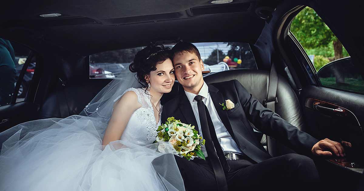 A bride and groom sit close together in the backseat of a car, smiling happily. The bride holds a bouquet of flowers and wears a white dress, while the groom is in a black suit with a boutonniere.