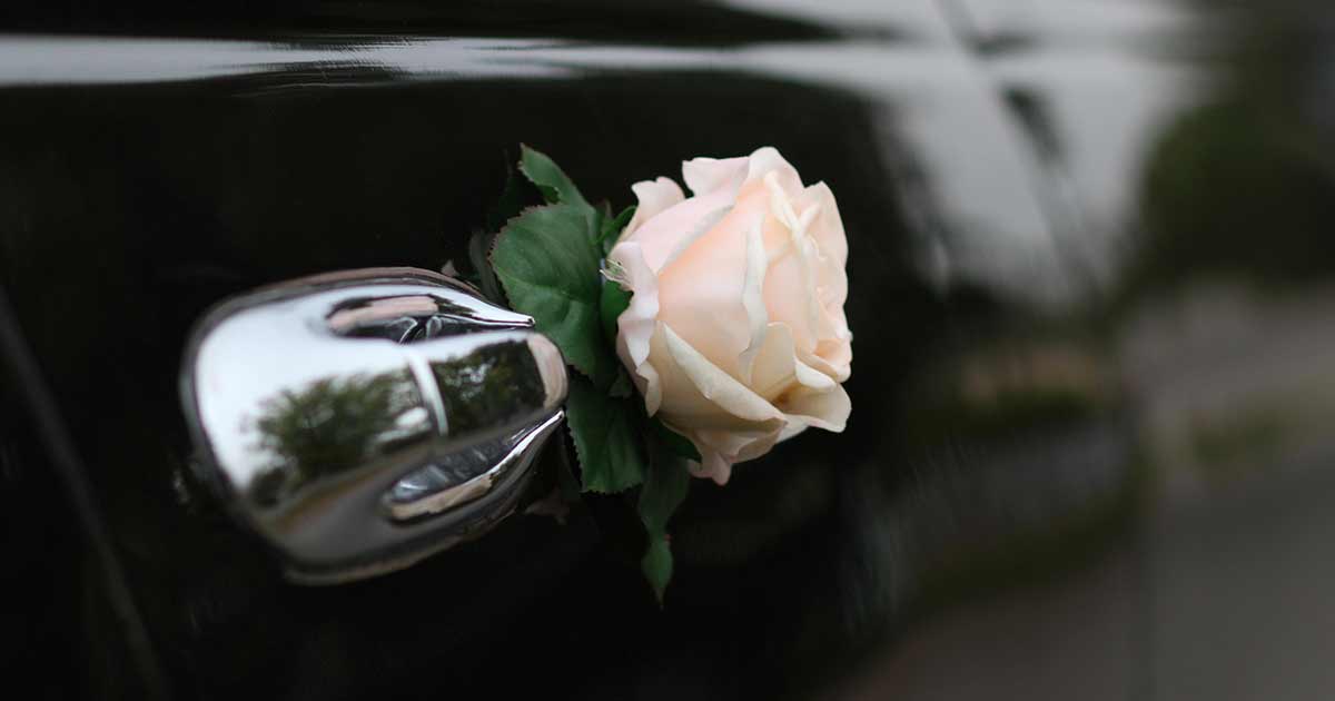 A pale pink rose is attached to the chrome door handle of a black car, with greenery around the flower. The background is blurred, highlighting the decoration.