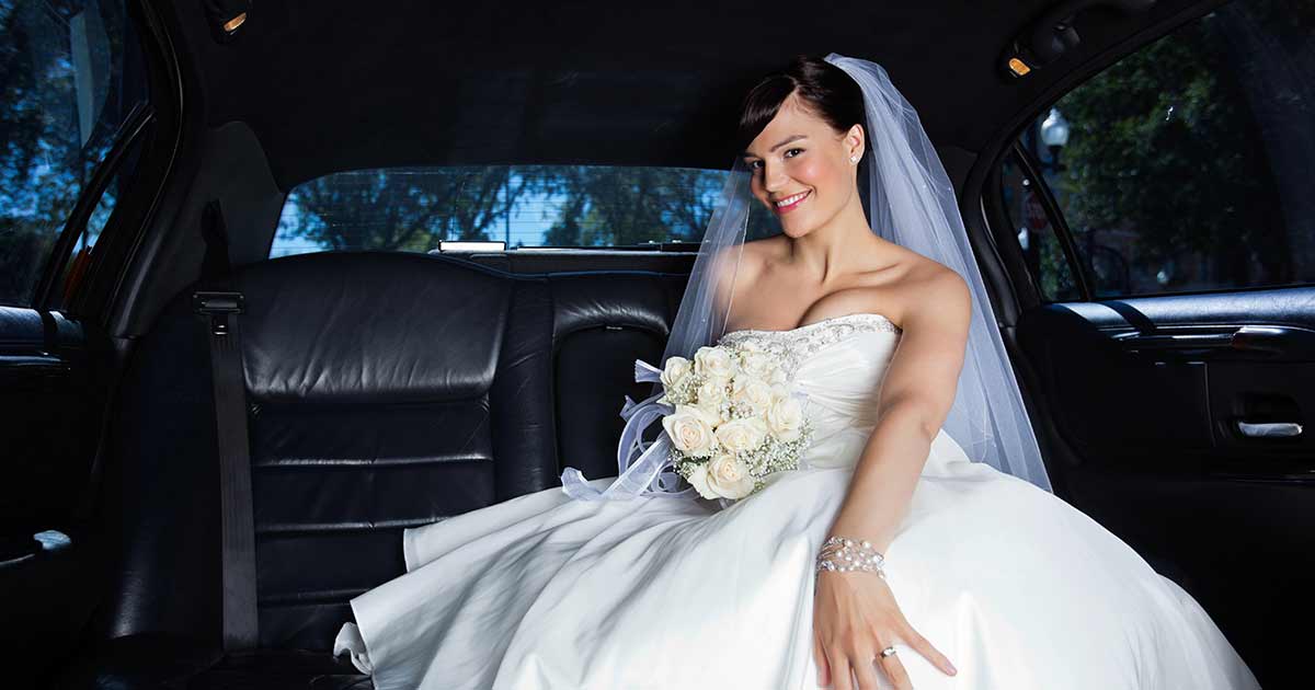 A bride in a white wedding dress and veil sits in the backseat of a car, smiling and showing her bouquet and ring.