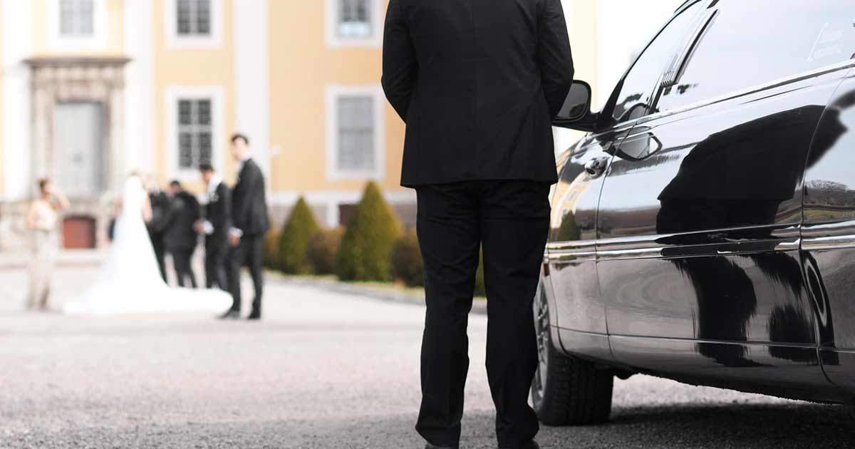 A person in a black suit stands near a black car, facing a wedding party blurred in the background outside a large, elegant building.
