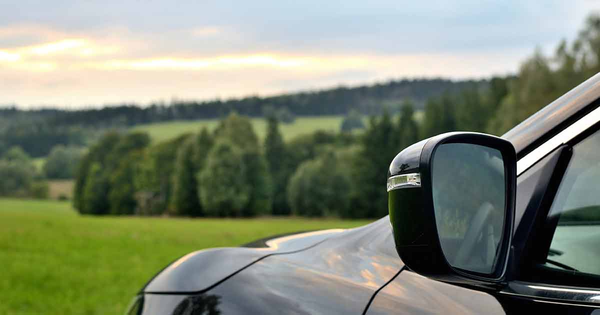 Close-up of a black car’s side mirror with a blurred background of green fields, trees, and a cloudy sky at sunset.