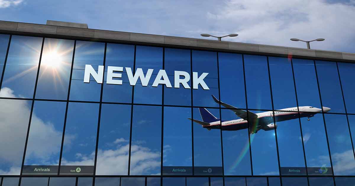 A large glass building with the word “NEWARK” on it reflects a jet airplane flying in a blue sky with clouds and sunlight.